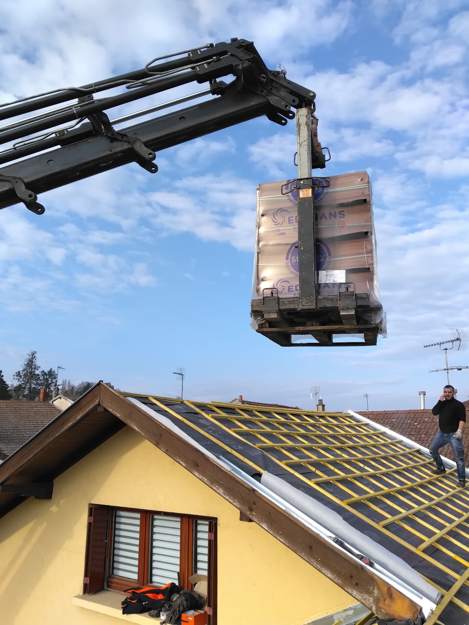 Rénovation de toiture : levage de tuiles Edilians par grue Grue de levage déposant une palette de tuiles Edilians sur une charpente de toit en cours de rénovation.