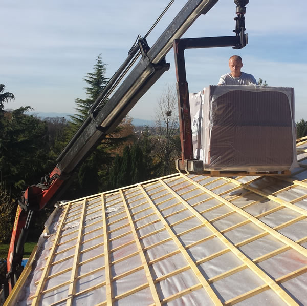 Livraison Matériaux de Toiture par Grue sur Chantier Grue levant une palette de tuiles sur une charpente de toit en bois en présence d'un couvreur.