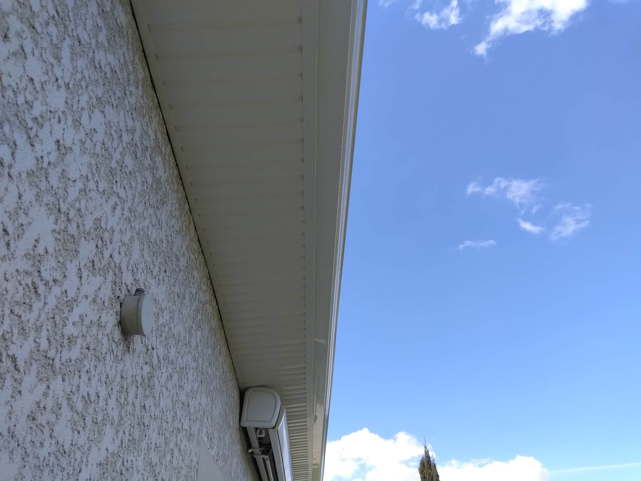 Finition de Toiture : Soffite et Gouttière sur Mur Crépi Détail d'un mur en crépi blanc, soffite et gouttière sous un ciel bleu dégagé avec quelques nuages.
