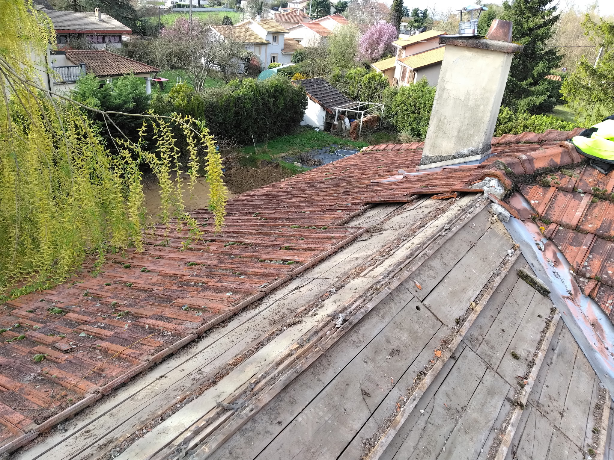 Rénovation de toiture : réparation de tuiles en terre cuite Toit en rénovation avec tuiles rouges manquantes révélant les lattes en bois et cheminée grise, vue plongeante.